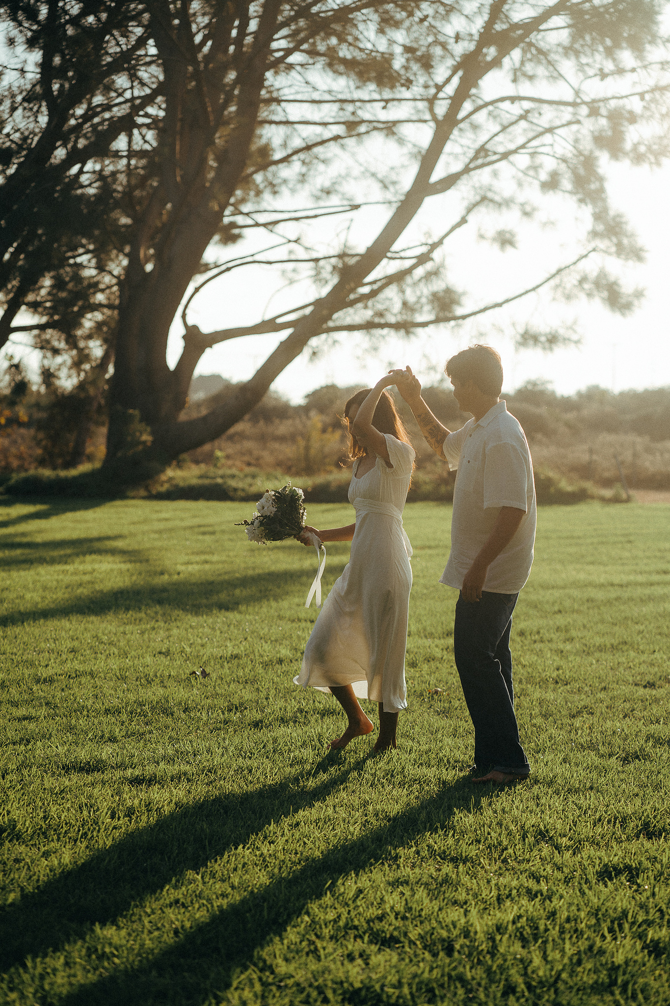 Two young lovebirds pose in a grassy field, the bride in a cottage-core white dress, in the afternoon sun for their engagement photo session in Fairview Park, Costa Mesa, CA. Engagement photography by Aly Rose Photo. California wedding photographer
