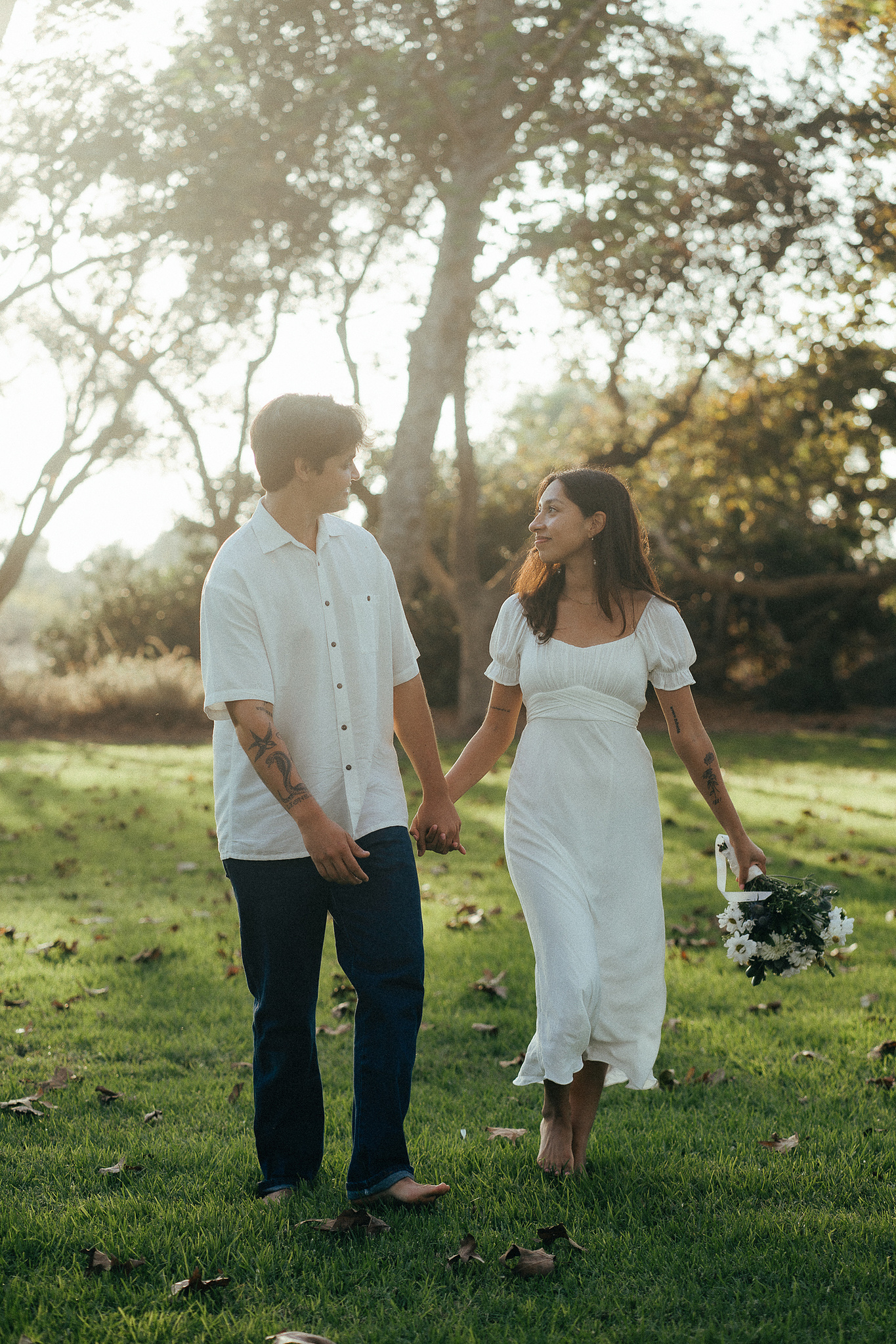 Two young lovebirds pose in a grassy tree grove, the bride in a cottage-core white dress, in the afternoon sun for their engagement photo session in Fairview Park, Costa Mesa, CA. Engagement photography by Aly Rose Photo. California wedding photographer