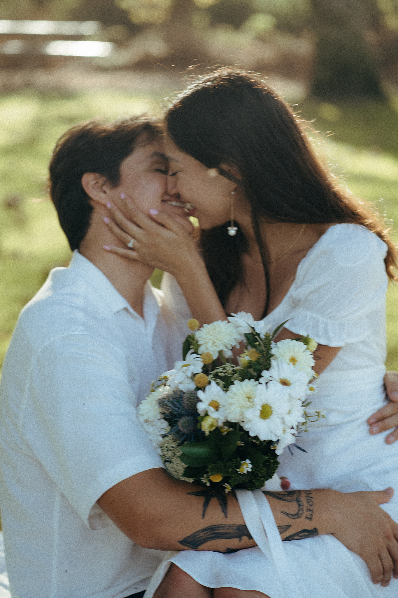 Two young lovebirds share a blurry, smiling kiss, the bride in a cottage-core white dress, in the afternoon sun for their engagement photo session in Fairview Park, Costa Mesa, CA. Engagement photography by Aly Rose Photo. California wedding photographer