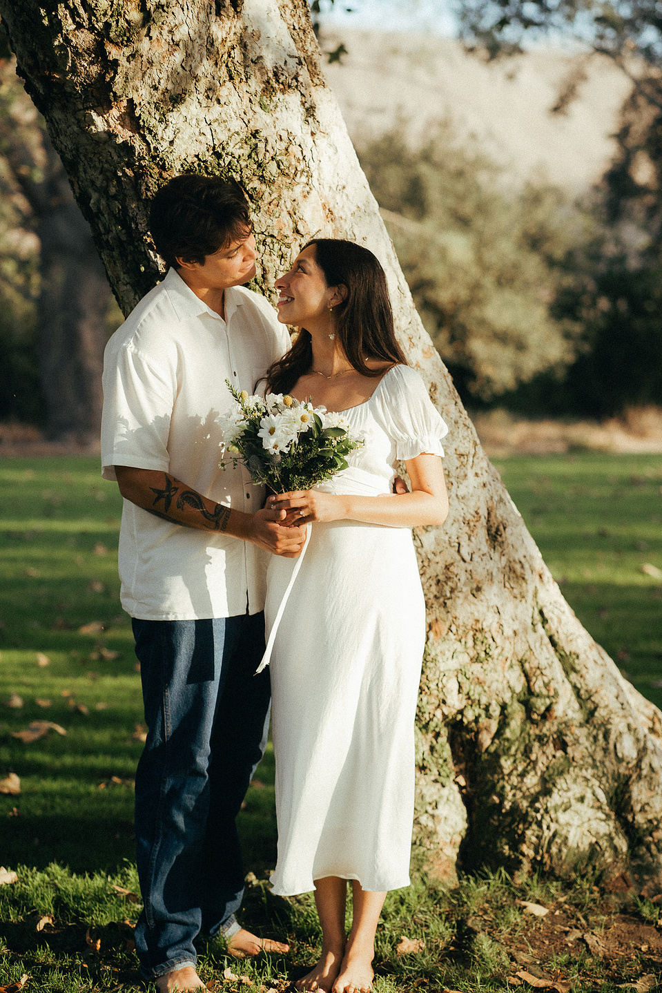 Two young lovebirds pose in front of a tree a cottage-core white dress and dress shirt in the afternoon sun for their engagement photo session in Fairview Park, Costa Mesa, CA. Engagement photography by Aly Rose Photo. California wedding photographer