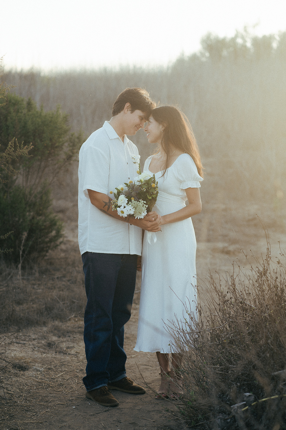 Two young lovebirds pose in a cottage-core white dress and dress shirt at golden hour for their engagement photo session in Fairview Park, Costa Mesa, CA. Natural light engagement photography by Aly Rose Photo. California wedding photographer