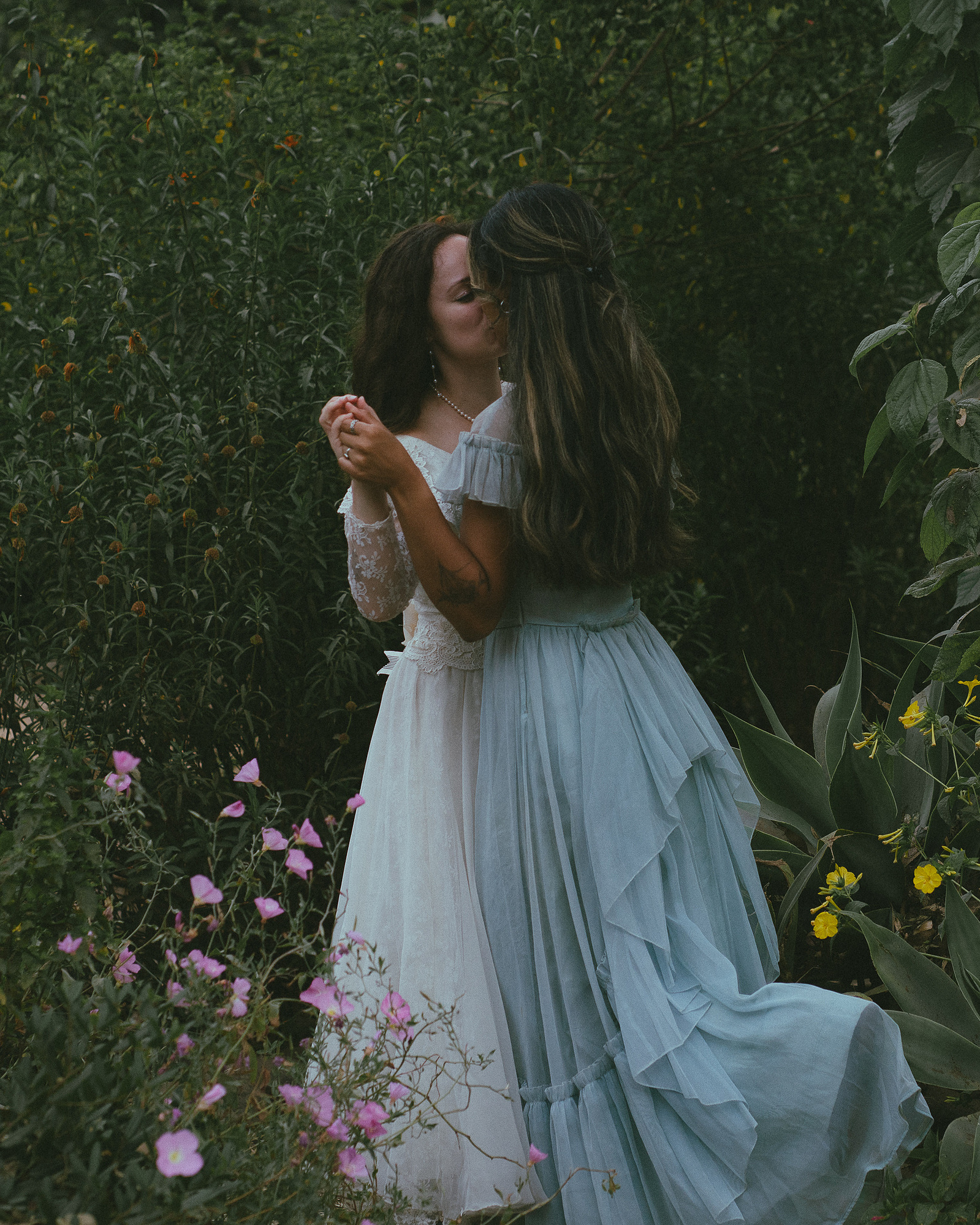Romantic, cottagecore, fairytale, LGBTQ portrait session in San Juan Capistrano, Orange County, CA. A woman in a blue dress kisses the hand of a woman in a white dress holding wildflowers. Photography by Aly Rose Photo, CA wedding, elopement photographer