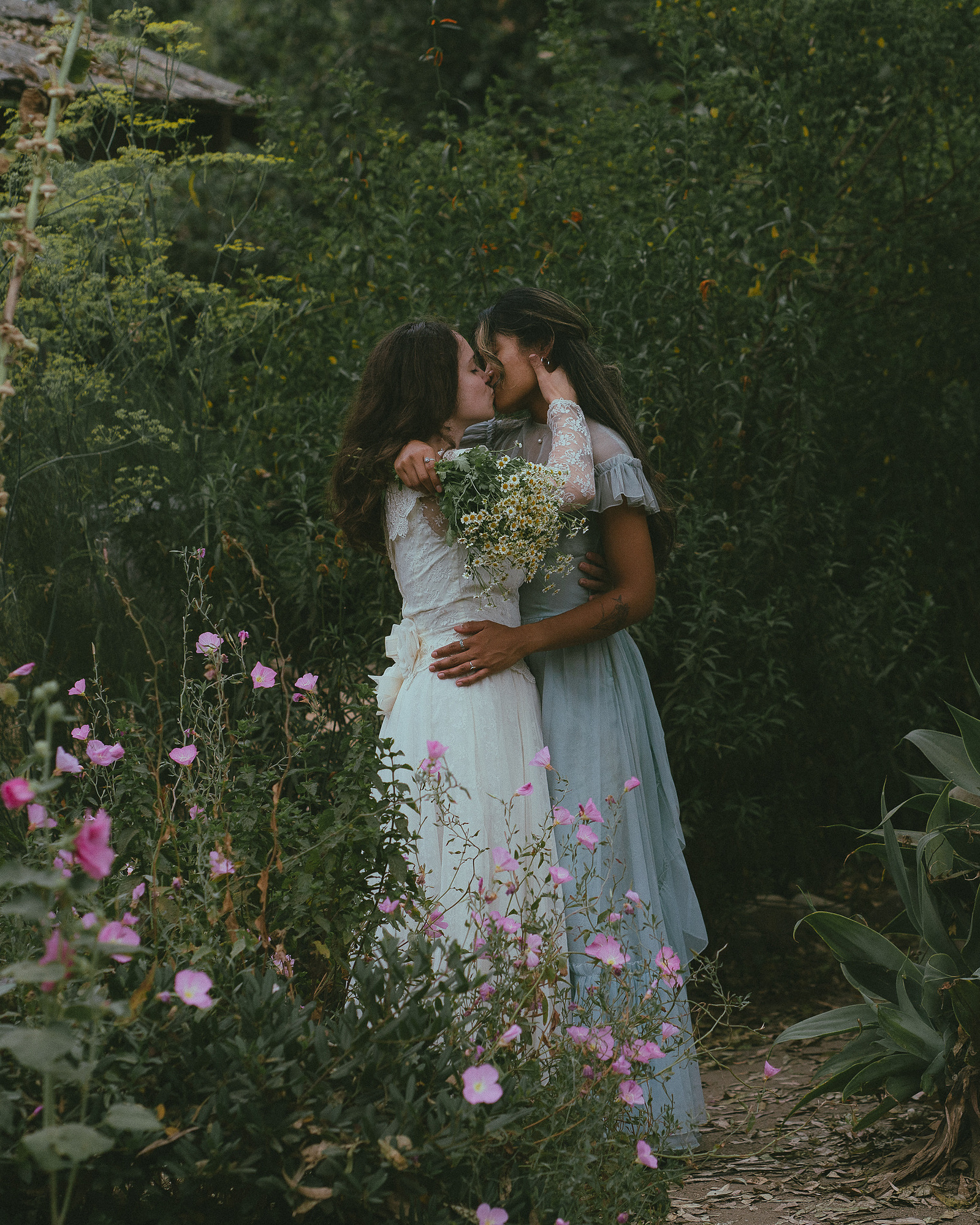 Romantic, cottagecore, fairytale, LGBTQ portrait session in San Juan Capistrano, Orange County, CA. A woman in a blue dress kisses the hand of a woman in a white dress holding wildflowers. Photography by Aly Rose Photo, CA wedding, elopement photographer