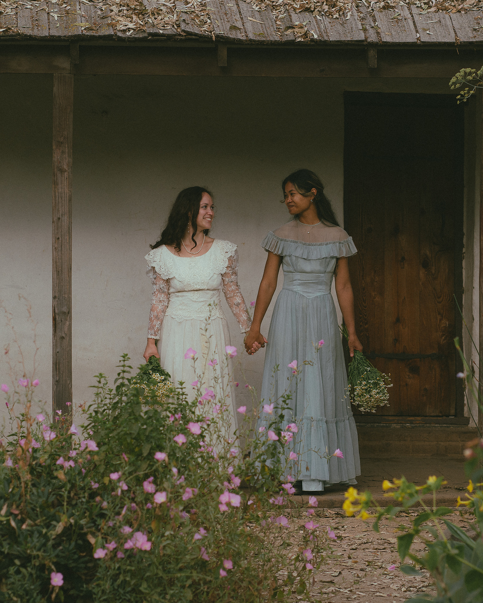 Romantic, cottagecore, fairytale, LGBTQ portrait session in San Juan Capistrano, Orange County, CA. A woman in a blue dress kisses the hand of a woman in a white dress holding wildflowers. Photography by Aly Rose Photo, CA wedding, elopement photographer
