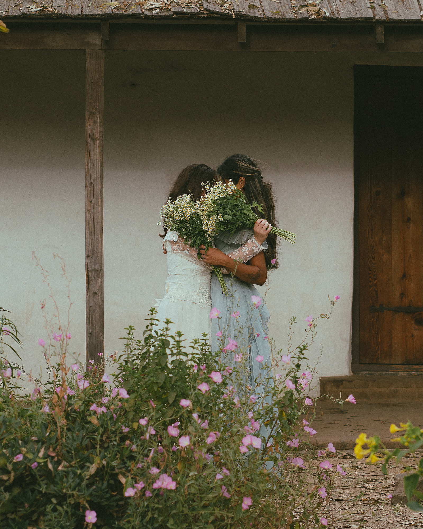 Romantic, cottagecore, fairytale, LGBTQ portrait session in San Juan Capistrano, Orange County, CA. A woman in a blue dress kisses the hand of a woman in a white dress holding wildflowers. Photography by Aly Rose Photo, CA wedding, elopement photographer