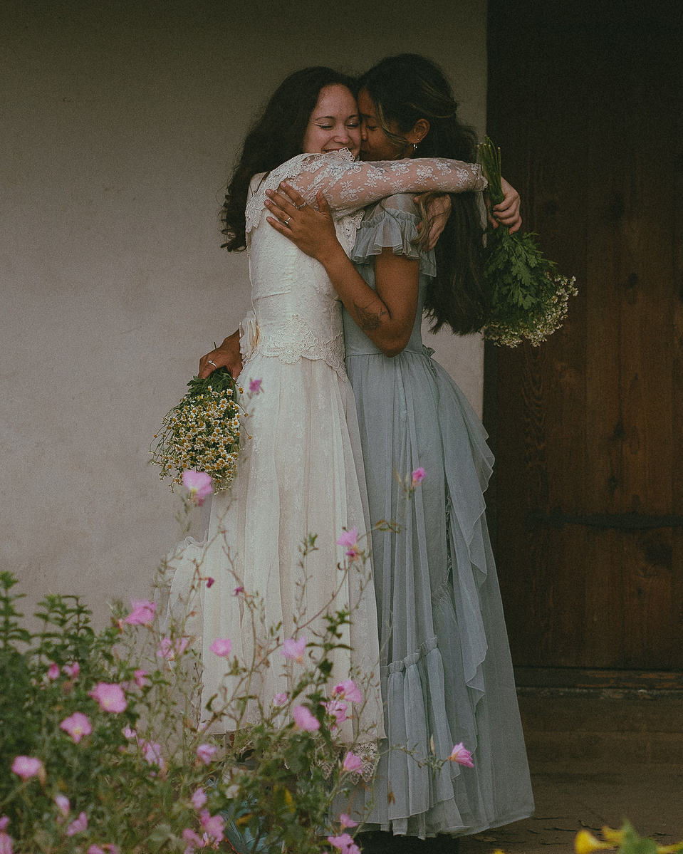 Romantic, cottagecore, fairytale, LGBTQ portrait session in San Juan Capistrano, Orange County, CA. A woman in a blue dress kisses the hand of a woman in a white dress holding wildflowers. Photography by Aly Rose Photo, CA wedding, elopement photographer