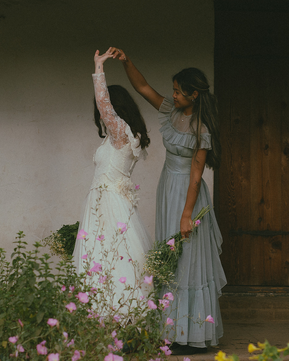 Romantic, cottagecore, fairytale, LGBTQ portrait session in San Juan Capistrano, Orange County, CA. A woman in a blue dress kisses the hand of a woman in a white dress holding wildflowers. Photography by Aly Rose Photo, CA wedding, elopement photographer