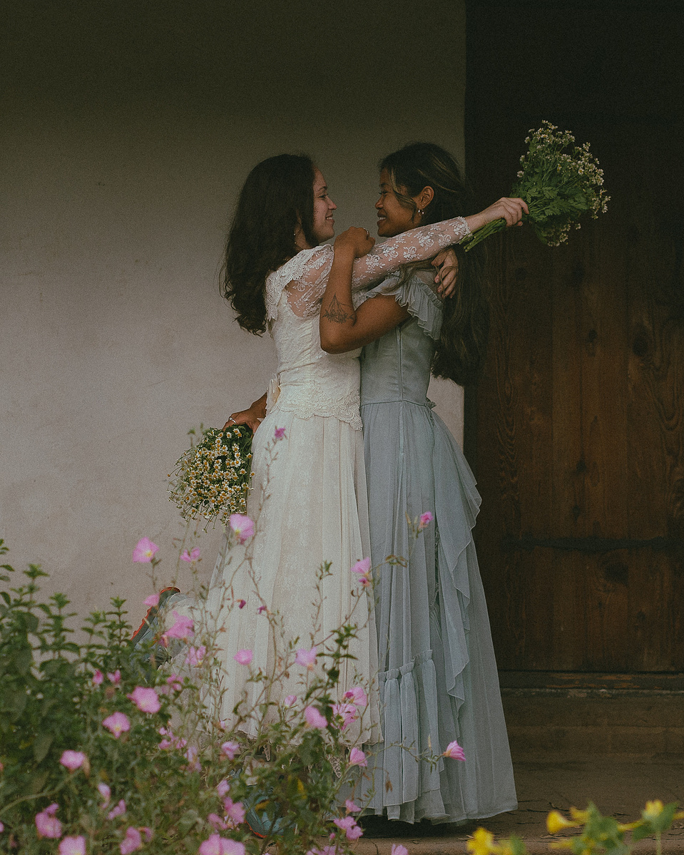 Romantic, cottagecore, fairytale, LGBTQ portrait session in San Juan Capistrano, Orange County, CA. A woman in a blue dress kisses the hand of a woman in a white dress holding wildflowers. Photography by Aly Rose Photo, CA wedding, elopement photographer