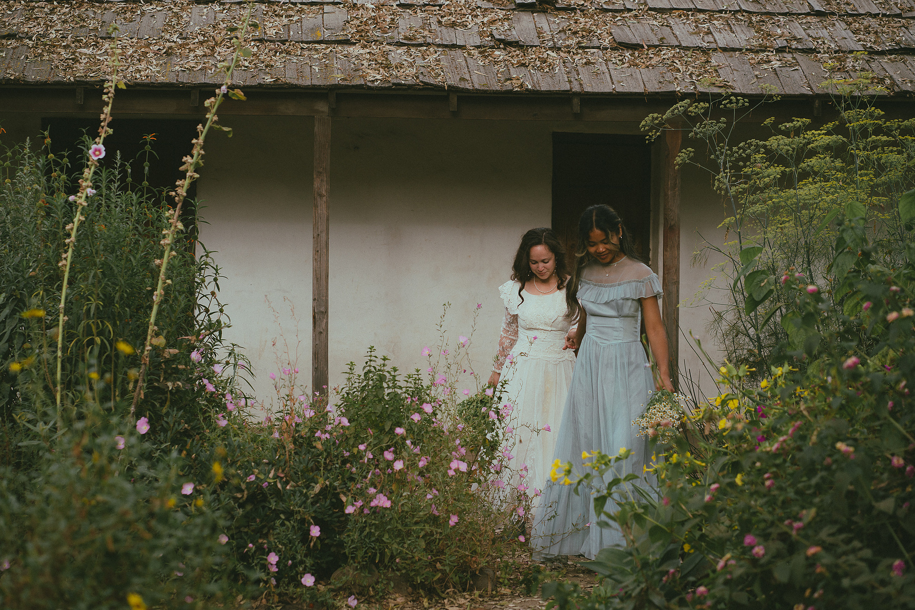 Romantic, cottagecore, fairytale, LGBTQ portrait session in San Juan Capistrano, Orange County, CA. A woman in a blue dress kisses the hand of a woman in a white dress holding wildflowers. Photography by Aly Rose Photo, CA wedding, elopement photographer