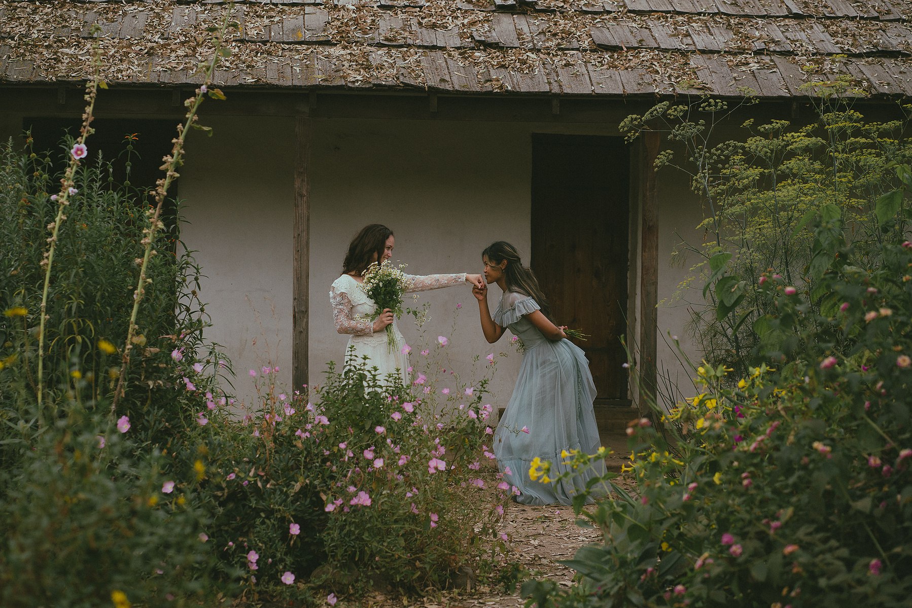 Romantic, cottagecore, fairytale, LGBTQ portrait session in San Juan Capistrano, Orange County, CA. A woman in a blue dress kisses the hand of a woman in a white dress holding wildflowers. Photography by Aly Rose Photo, CA wedding, elopement photographer