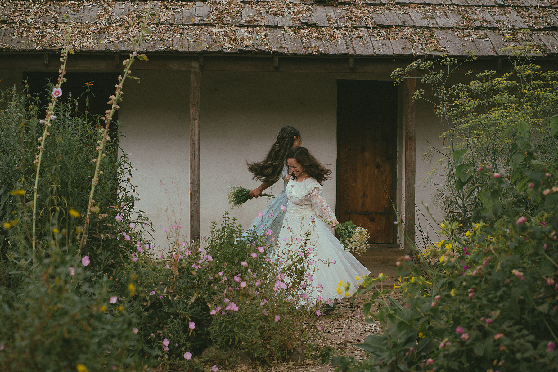 Romantic, cottagecore, fairytale, LGBTQ portrait session in San Juan Capistrano, Orange County, CA. A woman in a blue dress kisses the hand of a woman in a white dress holding wildflowers. Photography by Aly Rose Photo, CA wedding, elopement photographer