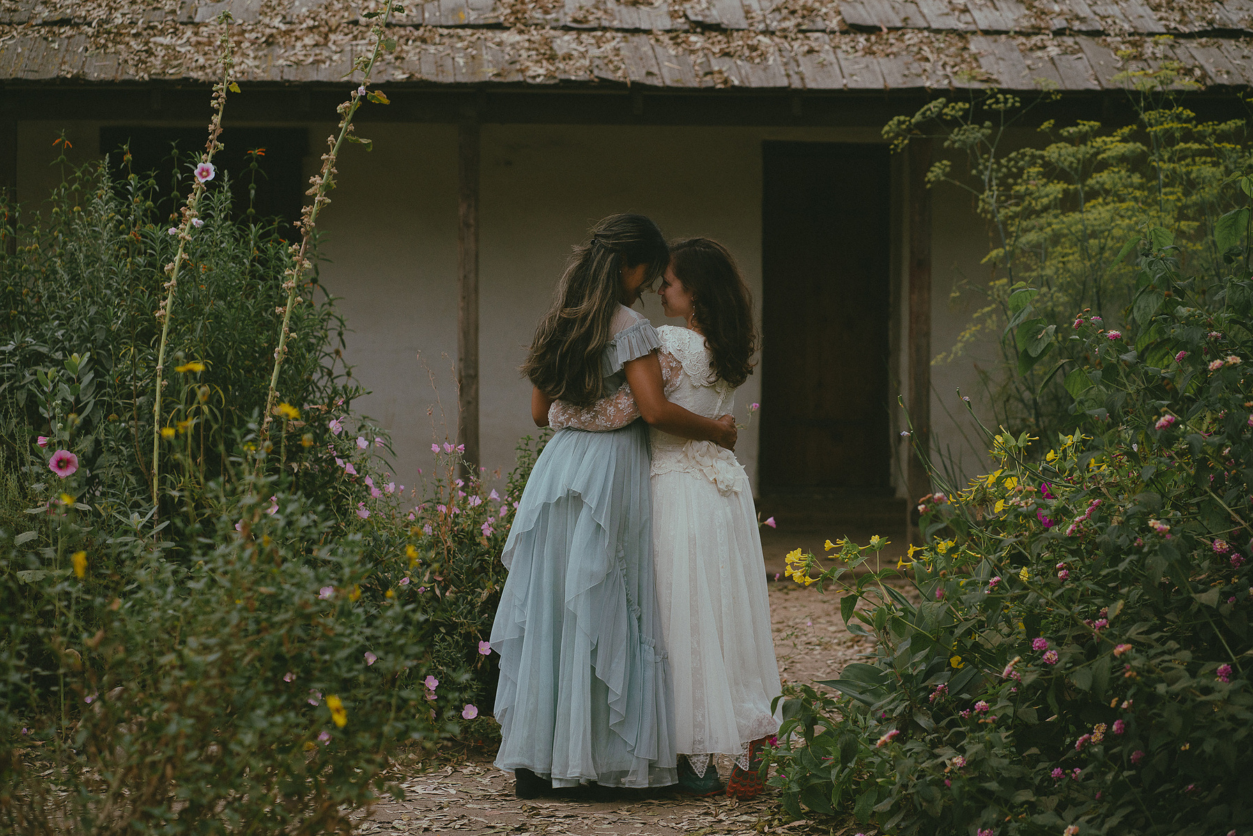 Romantic, cottagecore, fairytale, LGBTQ portrait session in San Juan Capistrano, Orange County, CA. A woman in a blue dress kisses the hand of a woman in a white dress holding wildflowers. Photography by Aly Rose Photo, CA wedding, elopement photographer