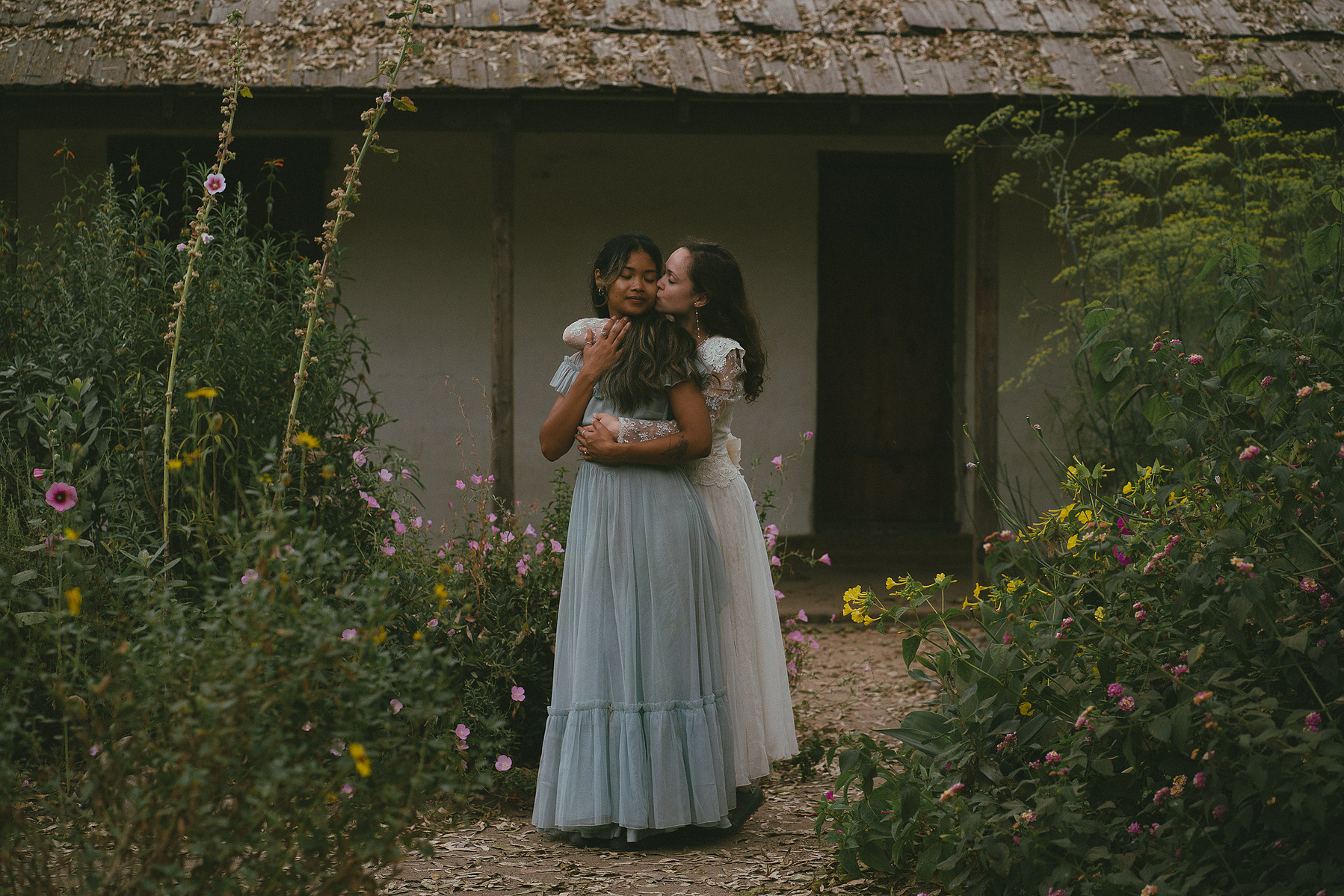 Romantic, cottagecore, fairytale, LGBTQ portrait session in San Juan Capistrano, Orange County, CA. A woman in a blue dress kisses the hand of a woman in a white dress holding wildflowers. Photography by Aly Rose Photo, CA wedding, elopement photographer