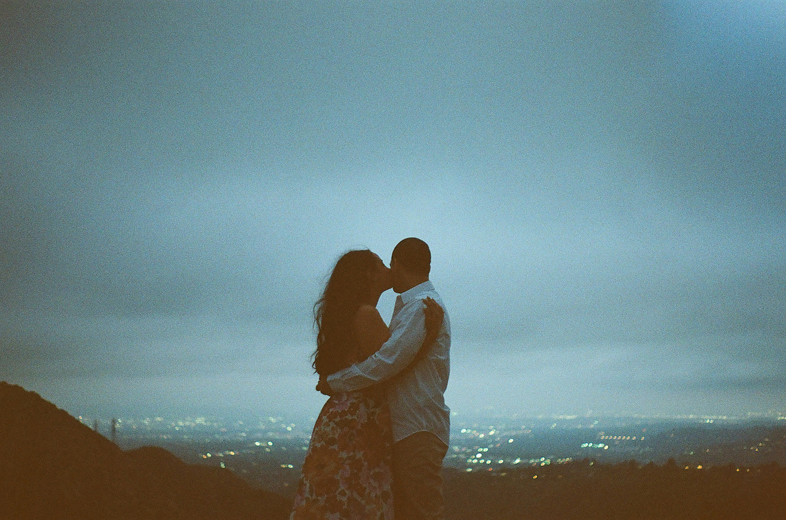 A cinematic engagement shoot up in the mountains with city lights twinkling below. Bride and groom to be embrace with a kiss. 35mm film photo. Romantic, dreamy vibes. Film and digital Southern California Wedding Elopement photographer Aly Rose Photo.  
