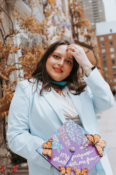 portrait of a nyu masters graduate student. she is a plus size woman with light skin and cool brown hair. She is wearing a light blue blazer and pants set and she's holding her purple grad cap that is decorated with monarch butterflies. 