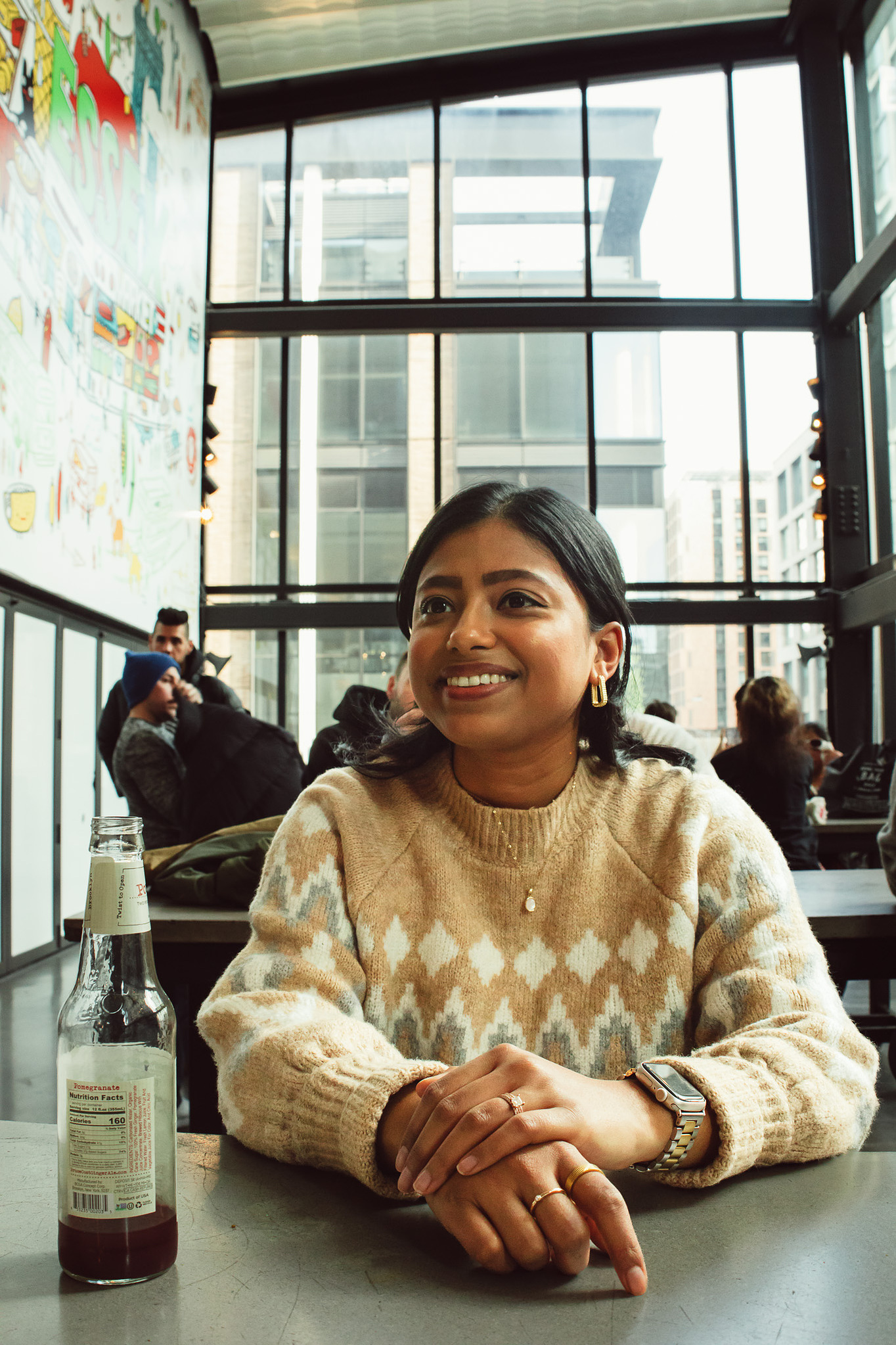 A young South Asian woman is wearing a patterned sweater and is smiling with her hands on the table. In the background you can see floor to ceiling windows of Essex Market in the Lower East Side.
