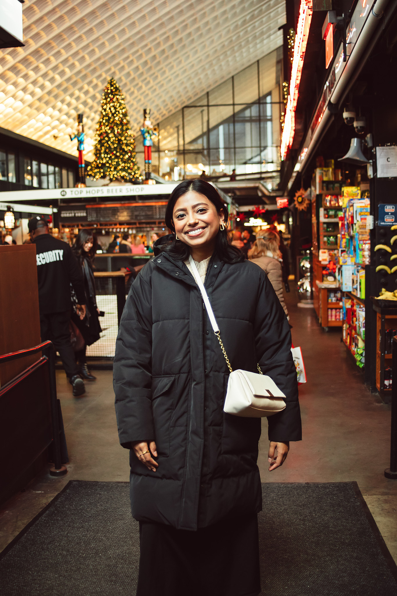 A South Asian young woman wearing a black long puffer coat inside of Essex Market in the Lower East Side of Manhattan. 
