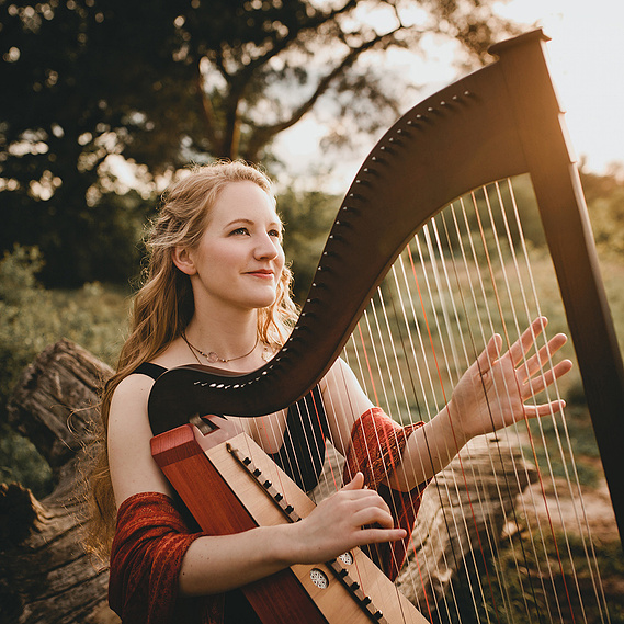 Eine Musikerin sitzt für ihr Künstlerportrait in der Natur mit ihrer Harfe in der Abendsonne und blickt verträumt in die Ferne.