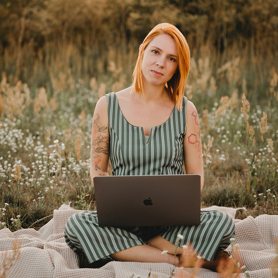 Eine Unternehmerin im gestreiften Jumpsuit sitzt beim Business Fotoshooting in der Natur mit ihrem Macbook auf einer Decke und blickt freundlich in die Kamera.