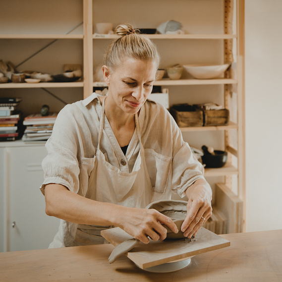Eine Keramikerin arbeitet während eines Business-Fotoshootings in ihrer Töpferwerkstatt lächelnd an einer Schale.