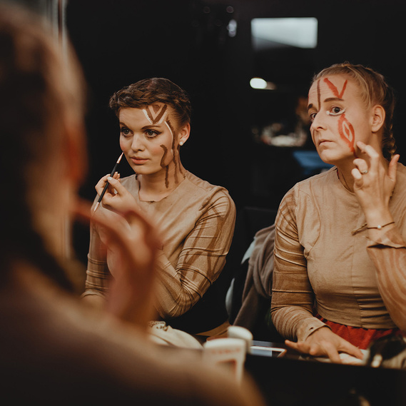 Zwei Darstellerinnen eines Musicals schminken sich in Vorbereitung auf die Generalprobe vor einem großen Spiegel.