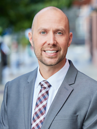 Gentlemen, with bald head, low scruffy beard, smiling for a professional headshot on a environmental blurred backdrop, with a gray suit on.