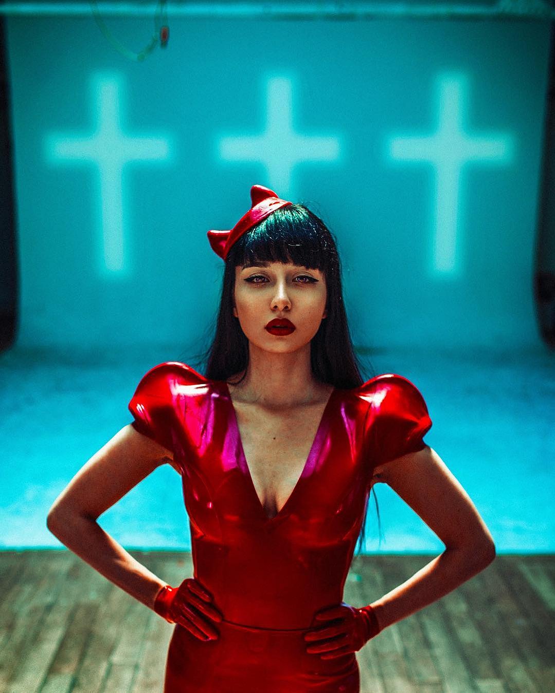 A woman in red latex stands in front of a projection of three crosses on the wall