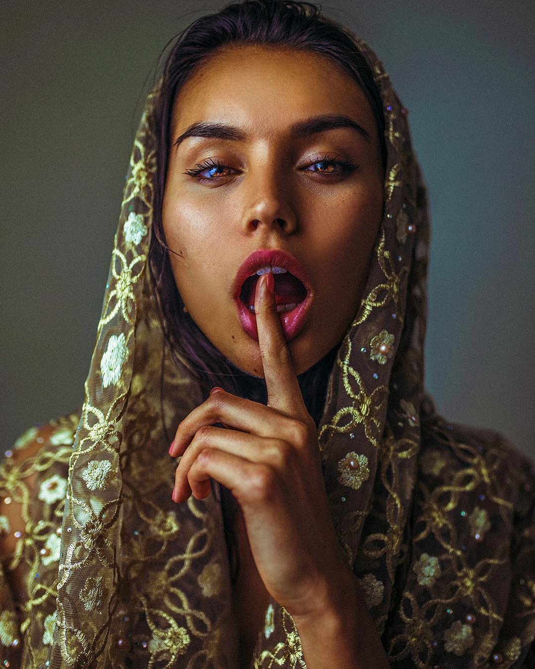 A woman with a simple scarf draped over her head, motions with a finger over her mouth for silence.