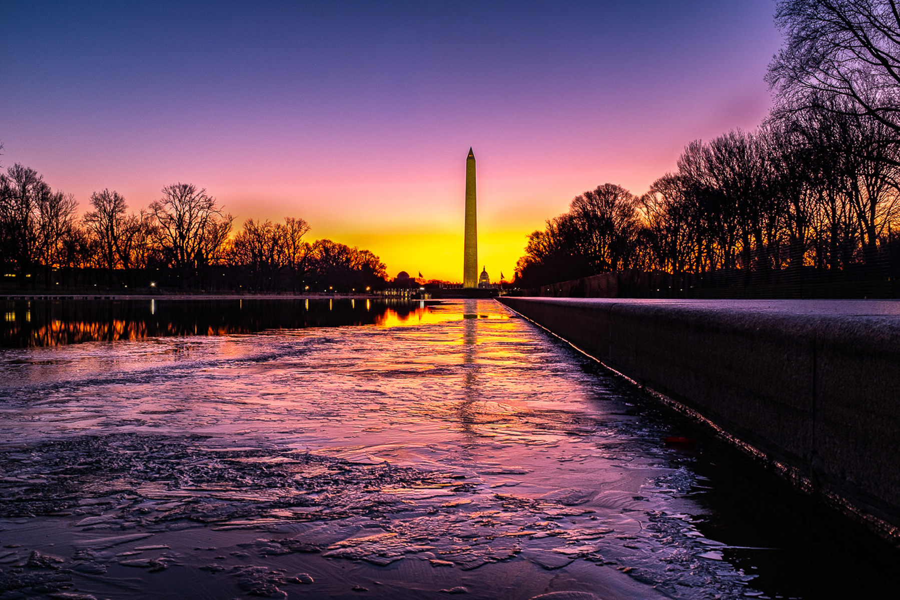 Winter Sunrise view of Lincoln Memorial Reflecting Pool with Washington Monument in the distance, showing pink and yellow sunrise lights reflected on the ice on the pool, and leading lines from deck of pool on the right side of image. 