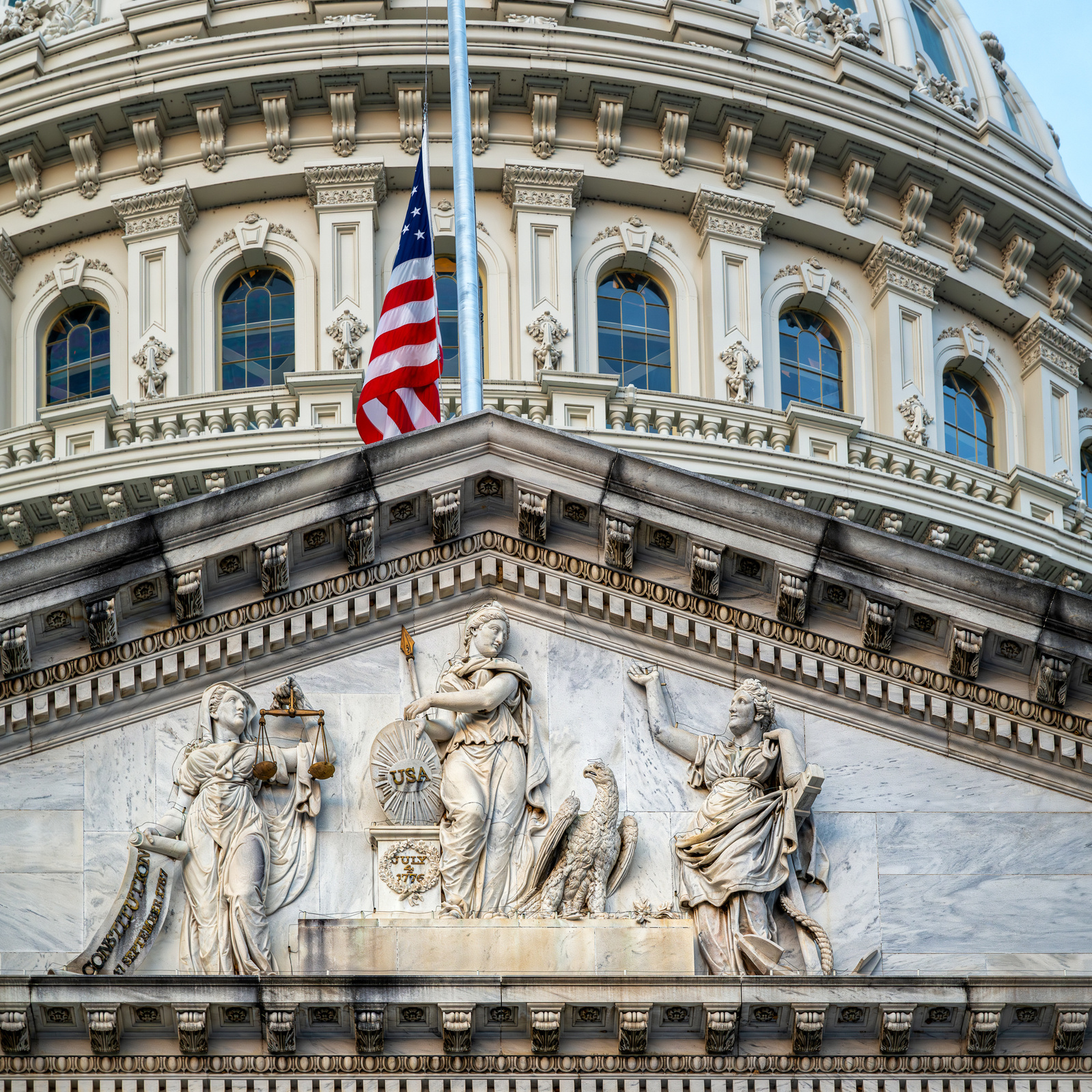Pediment just below dome of U.S. Capitol showing "Genius of America" statue grouping, east side of Capitol, with U.S. Flag at half staff  U.S. Flag at half-staff at its apex, with the three Genius figures (L-R: Justice, America and Hope) as its base. 