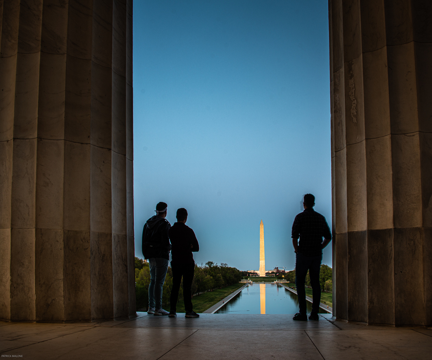 Silhouette of people looking out from Lincoln Memorial front pillars to the Washington Monument in the distance during blue hour. Pillars frame the people.