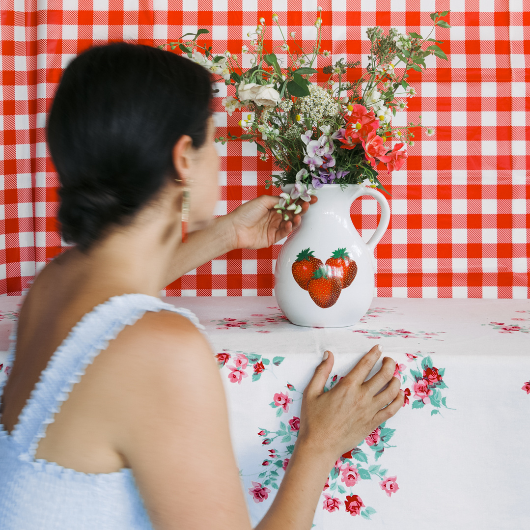 Artist in a blue dress building her scene of flowers in a white pitcher with strawberries on it.  Red and white gingham background and floral tablecloth.  