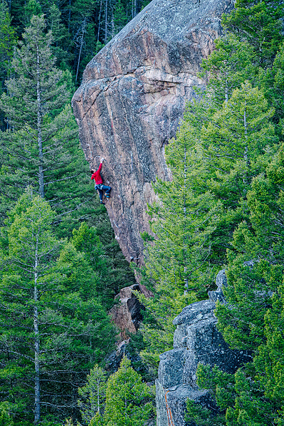 a person is rock climbing a steep and overhanging rock wall in a red sweatshirt. The rock type is granite gneiss located in the gallatin canyon of montana. The rock is surrounded by trees.