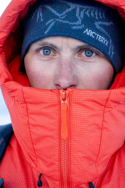 A man stand for a portrait while in the Alaska mountain cold air and wearing a red down puffy jacket.