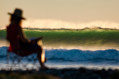 A women sits in a beach chair out of focus foreground with an ocean wave in the background that is in focus and slightly backlit. A peaceful scene.