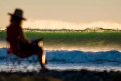 A women sits reading in a chair in a calm and warm tone image. The subject is out of focus and the background is of the ocean and a wave that is crashing down. 
