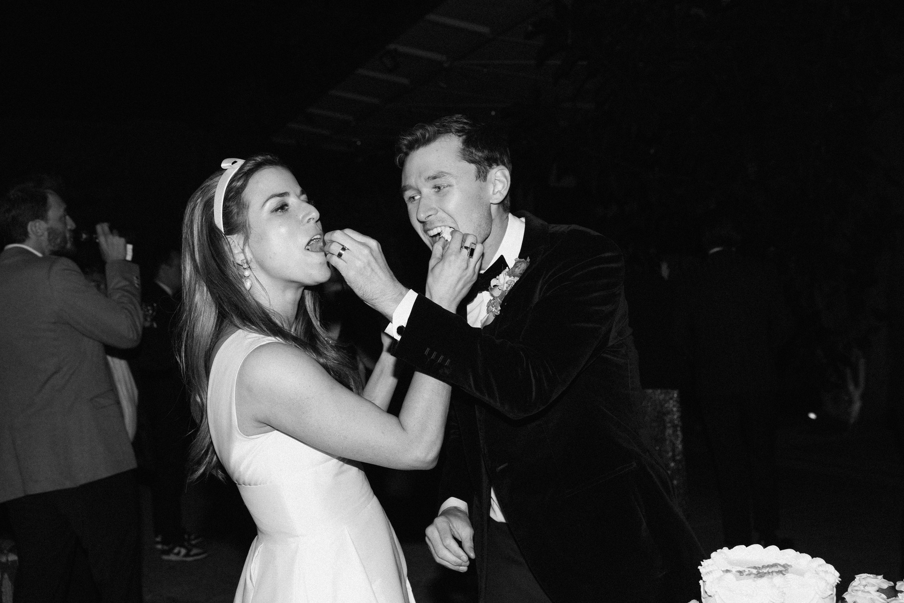 Black and white candid flash shot of bride and groom eating cake, captured by Abigail Gorden Photography. A playful and intimate wedding moment.