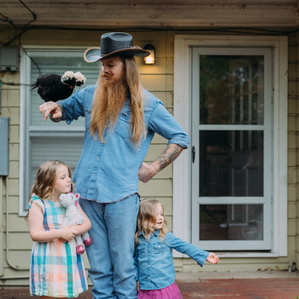 Father with beard and cowboy hat holding chicken while daughters stand next to him