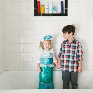 Little sister in mermaid costume holds hands with brother in tub