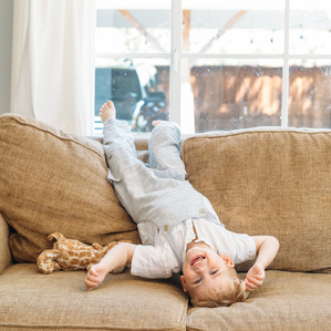 Little boy in overalls hangs upside down on couch laughing