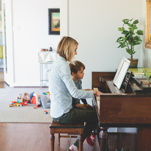 Mother and son sit together playing piano while other son plays with leggos