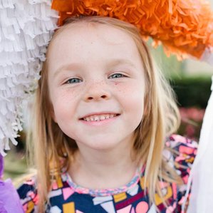 Girl with freckles stands with piñata on head smiling