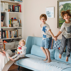 Two brothers jumping on couch in living room next to Mom and sister playing together and smiling