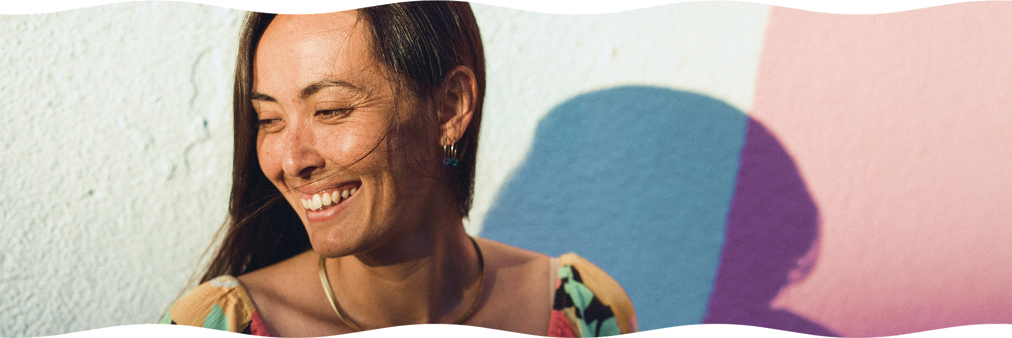 Sio Tepper smiles and looks to her right, a strand of hair crossing her face as her shadow is cast on the painted wall behind her by the waning, golden hour light, while the wavy edges of the image reflect Sio's ocean roots.