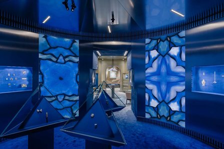 round room with metal walls and screens, pyramidal jewellery cases in front and champagne coloured room at the back