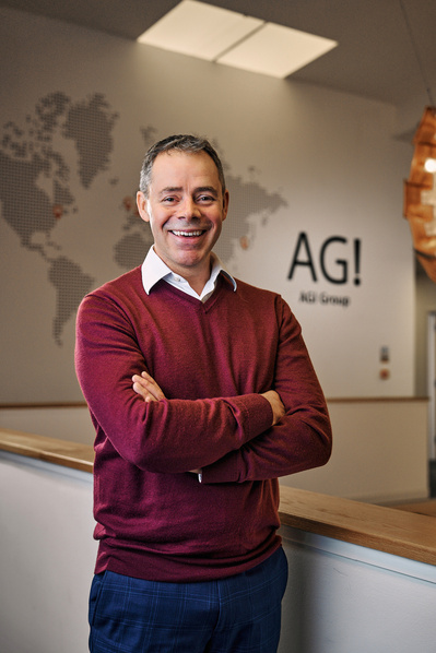 A portrait of a businessman stood in his office with his company logo on the wall behind him. 