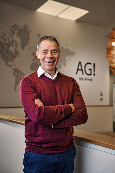 A portrait of a businessman stood in his office with his company logo on the wall behind him. 