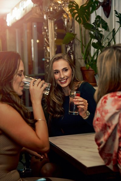 Three women enjoy a champagne sat at a table in a trendy bar. 