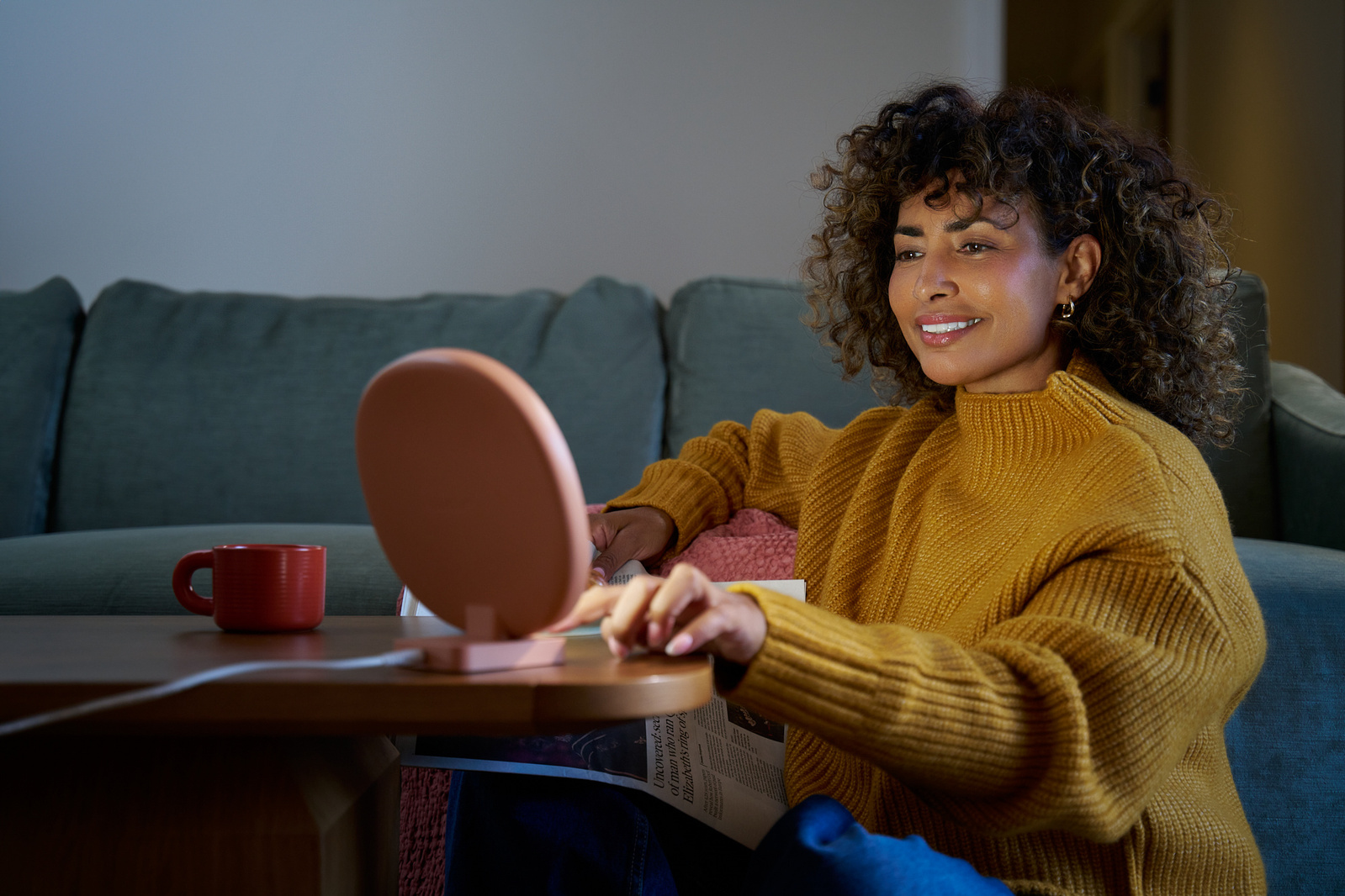 A stylish black woman with a cozy yellow jumper and afro hair is sitting on the floor and turning on her pink Lumie Lamp. 