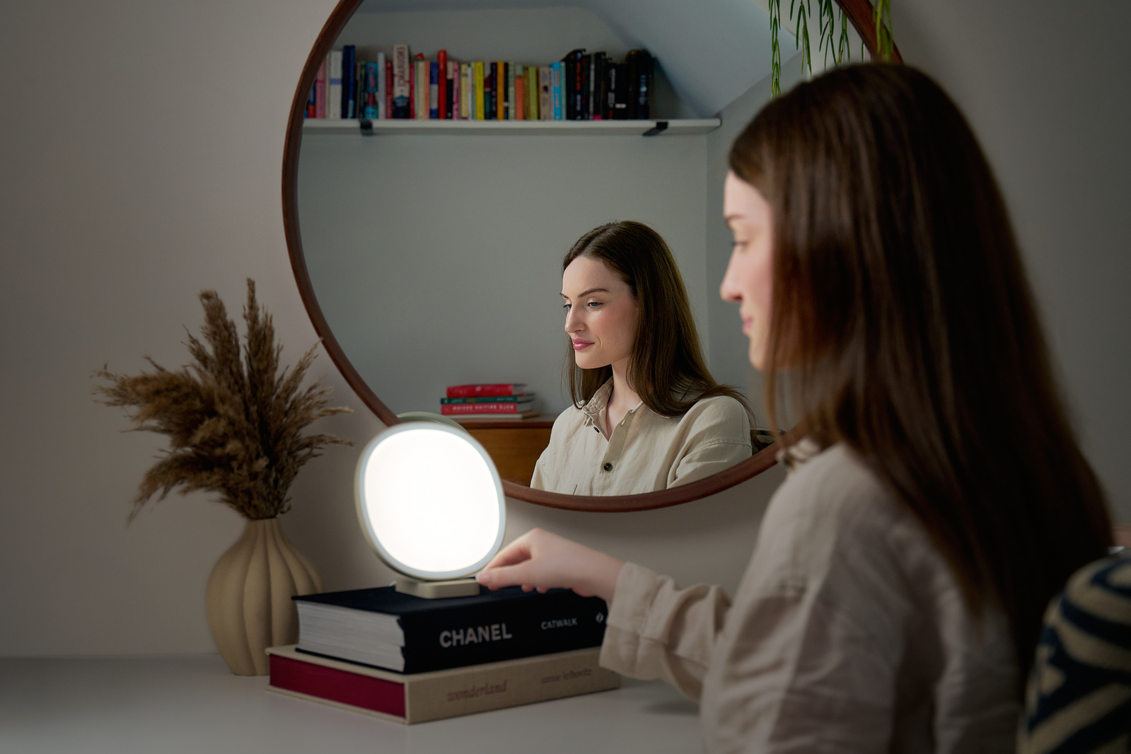 A woman sits at her large bedroom mirror with her Lumie lamp. 